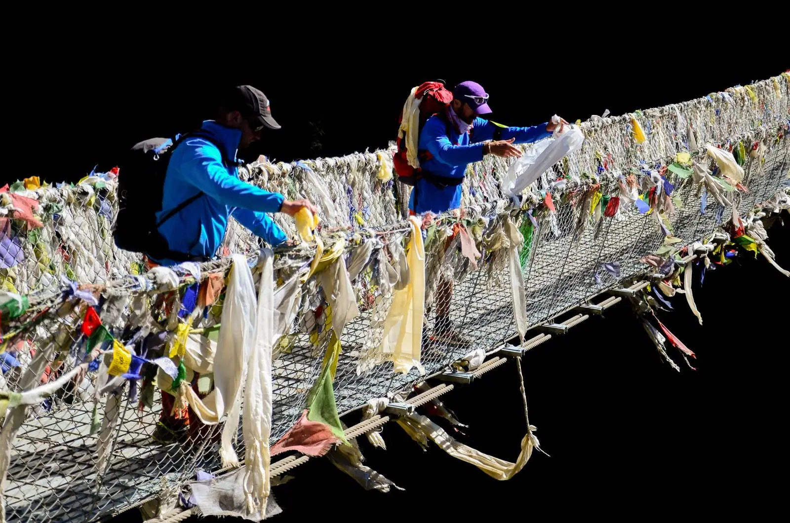Dos excursionistas atan pañuelos khata blancos a un puente colgante cubierto de banderas de oración.